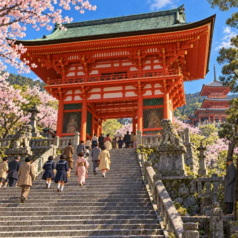 Visitors climb stone stairs toward a grand vermilion gate in Kyoto, with cherry blossoms, a hillside pagoda, and a man in a grey coat looking on.